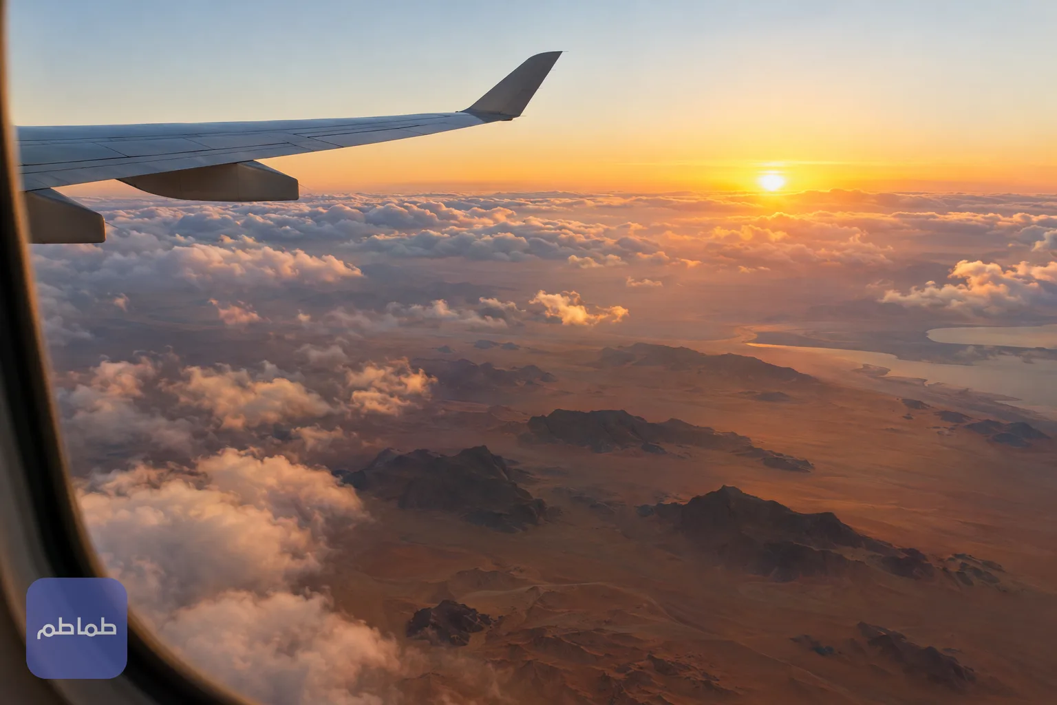 نافذة طائرة تطل على السحاب والأرض الصحراوية عند شروق الشمس