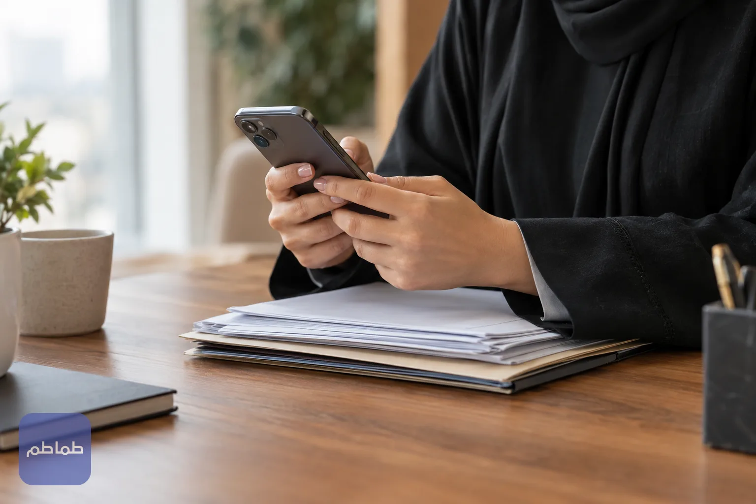 يد تمسك هاتفًا ذكيًا فوق مستندات مرتبة على مكتب خشبي، تعبيرًا عن تقديم الطلب إلكترونيًا